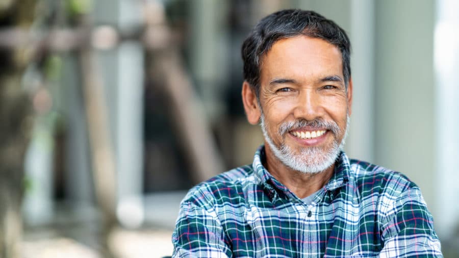 Older man with gray and white beard and plaid shirt smiling outside dental implants washington mi dentist